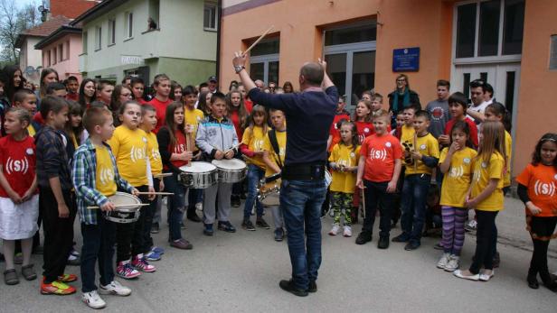 Eine Gruppe Kinder mit Trommeln wird von einem Mann dirigiert.