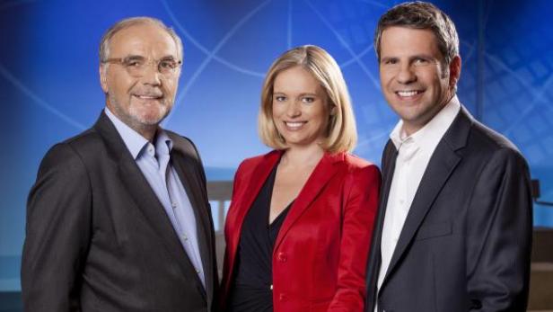 Drei Personen, vermutlich Nachrichtensprecher, stehen vor einem blauen Hintergrund.