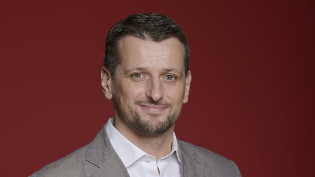 Ein Mann mit Bart und grauem Sakko lächelt vor einem roten Hintergrund.