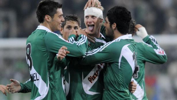 Eine Gruppe Fußballspieler in grünen Trikots umarmt einen Spieler mit einem Kopfverband.