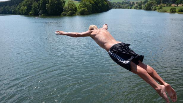 Ein junger Mann springt mit ausgebreiteten Armen in einen See.