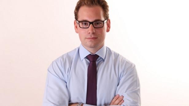 Ein Mann mit Brille, Hemd und Krawatte steht mit verschränkten Armen vor einem weißen Hintergrund.