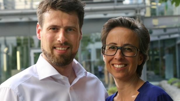 Ein Mann und eine Frau lächeln in die Kamera, im Hintergrund ein modernes Gebäude.