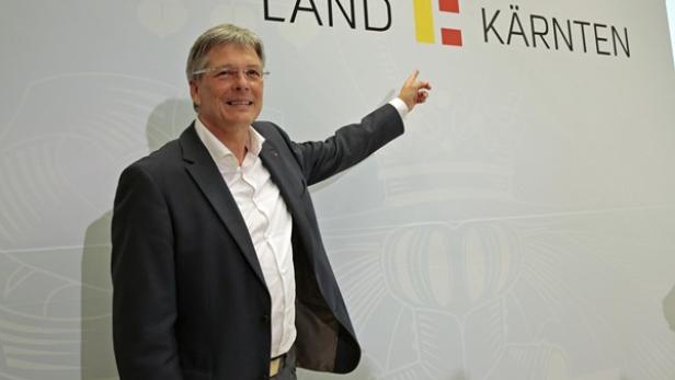 Ein Mann im Anzug zeigt auf ein Schild mit der Aufschrift „Land Kärnten“.