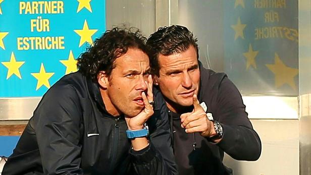 Zwei Männer sitzen nebeneinander, einer zeigt mit dem Finger nach vorne.