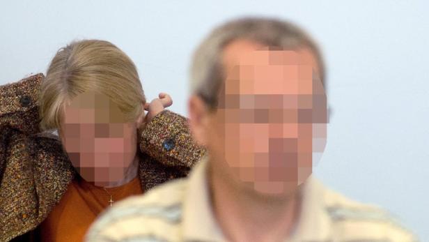 Eine Frau mit brauner Jacke und ein Mann stehen vor einer hellen Wand.