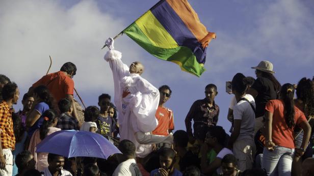 Ein Mann in weißer Kleidung schwenkt die Flagge von Mauritius inmitten einer Menschenmenge.
