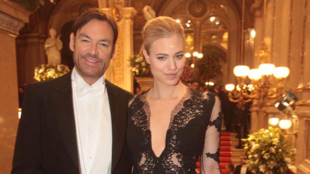 Ein Mann im Smoking und eine Frau in einem Spitzenkleid posieren in einem eleganten Ballsaal.