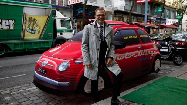 Ein Mann posiert vor einem roten Auto mit dem Aufdruck „Motor Village“ in einer städtischen Umgebung.
