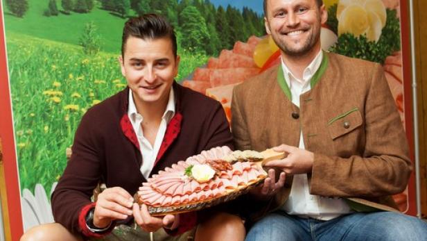 Zwei Männer in Tracht sitzen vor einem Schirnhofer-Banner und halten eine Wurstplatte.