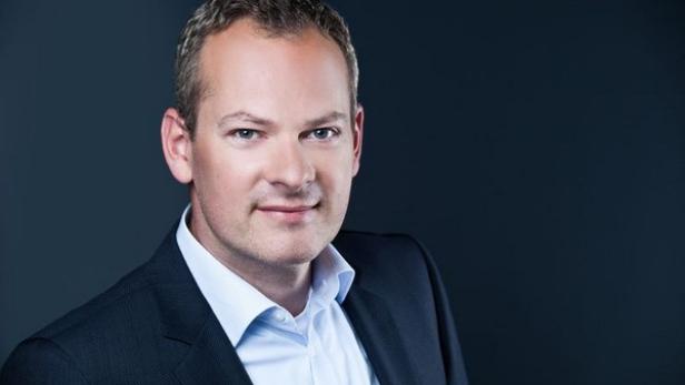 Ein Mann mit Anzug und hellblauem Hemd vor einem dunklen Hintergrund.