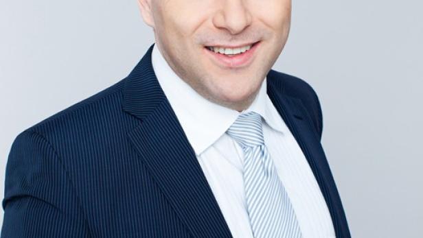 Ein lächelnder Mann im Anzug mit Krawatte vor einem grauen Hintergrund.