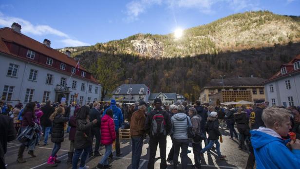 Eine Menschenmenge versammelt sich auf einem Platz vor einer bewaldeten Hügellandschaft.