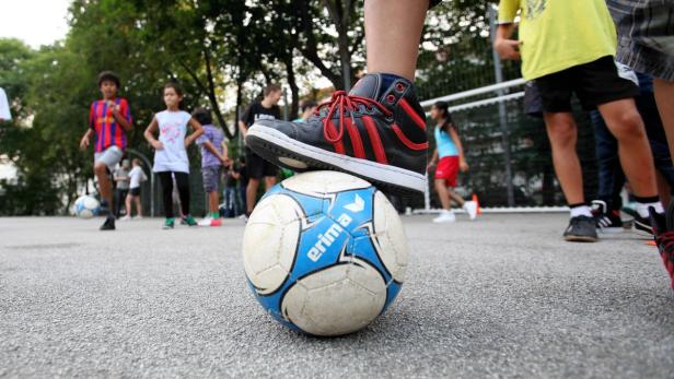 Ein Fuß in einem schwarzen Turnschuh steht auf einem Fußball, während Kinder im Hintergrund spielen.