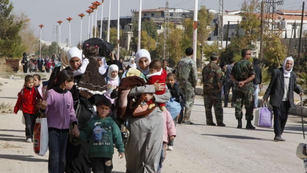 Eine Gruppe von Frauen und Kindern geht eine Straße entlang, begleitet von Soldaten.