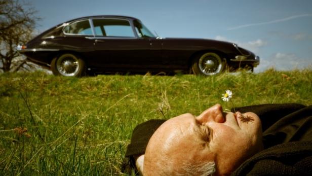 Ein Mann liegt im Gras mit einer Blume neben seinem Gesicht, im Hintergrund ein Jaguar E-Type.