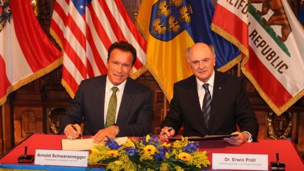 Arnold Schwarzenegger und Erwin Pröll unterzeichnen ein Dokument vor verschiedenen Flaggen.