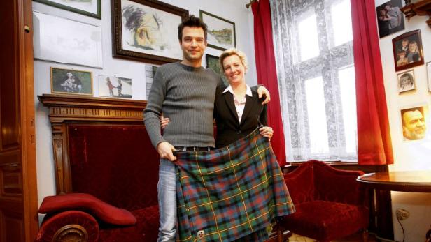 Ein Mann und eine Frau halten einen Schottenrock in einem Wohnzimmer hoch.