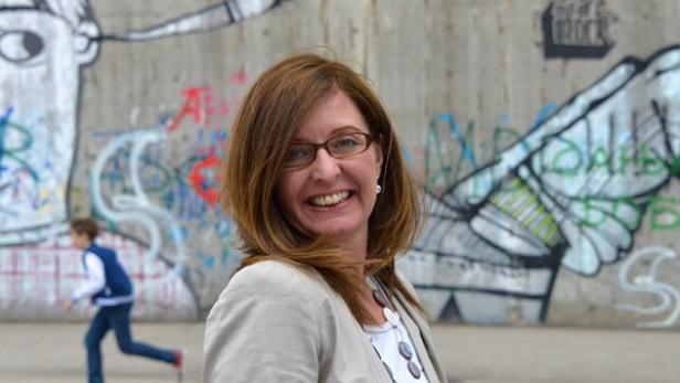 Eine lächelnde Frau mit Brille posiert vor einer Wand mit Graffiti, während ein Junge im Hintergrund rennt.
