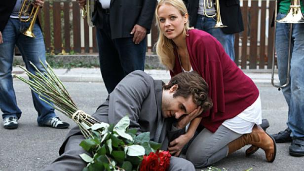 Eine Frau kniet neben einem am Boden liegenden Mann, umgeben von Musikern und Blumen.