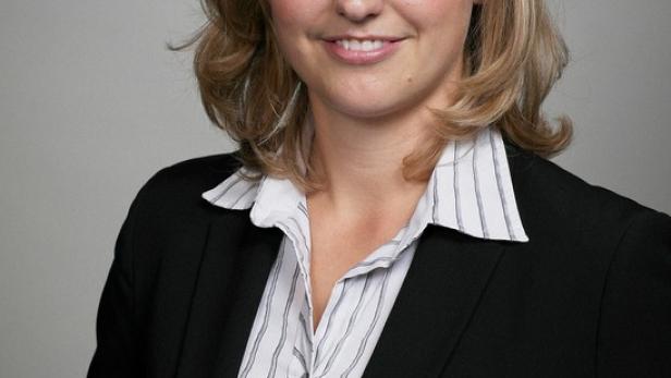 Eine blonde Frau in einem schwarzen Blazer und einer gestreiften Bluse lächelt.