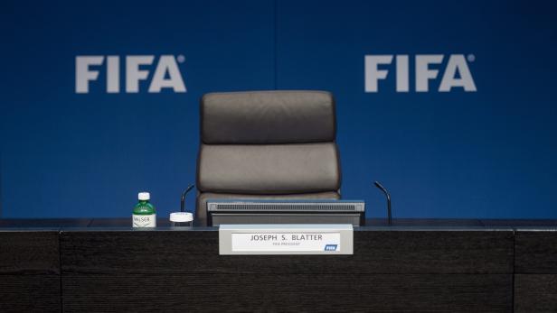 Ein leerer Stuhl mit dem Namensschild „Joseph S. Blatter, FIFA Präsident“ vor einem FIFA-Hintergrund.