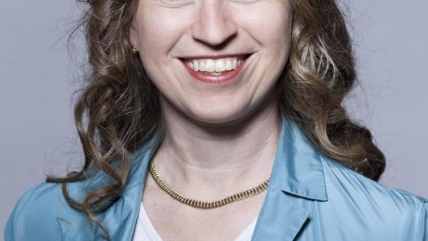 Eine lächelnde Frau mit lockigem Haar, gekleidet in eine blaue Jacke und eine goldene Halskette.