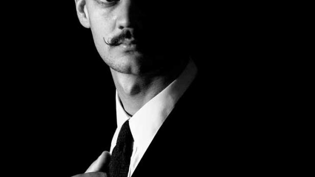 Ein Mann mit Schnurrbart im Anzug, zur Hälfte im Schatten.