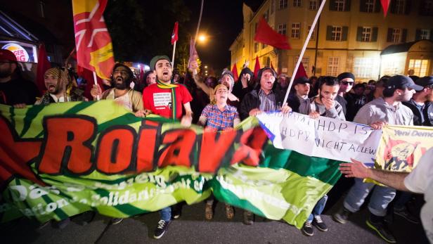 Eine Demonstration mit Menschen, die Fahnen und ein Banner mit der Aufschrift „Rojava“ tragen.