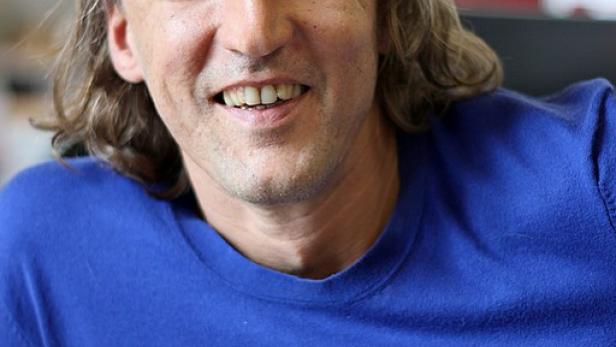 Ein lächelnder Mann mit langen blonden Haaren und einem blauen Pullover.