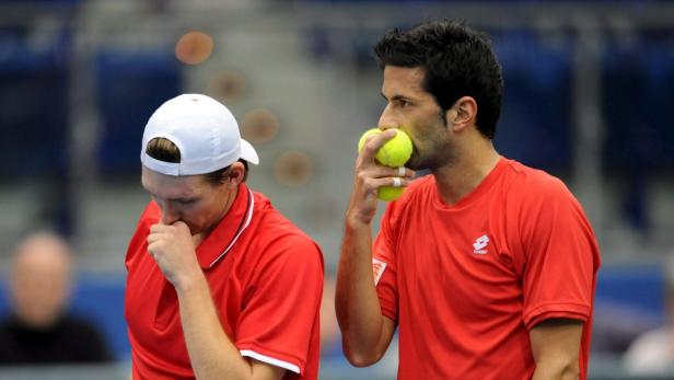 Zwei Tennisspieler in roten Trikots wirken enttäuscht; einer hält Tennisbälle im Mund.