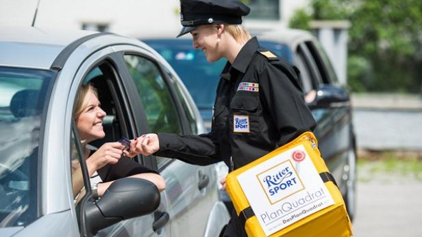 Eine Frau im Auto nimmt von einer uniformierten Person eine Tafel Ritter Sport entgegen.