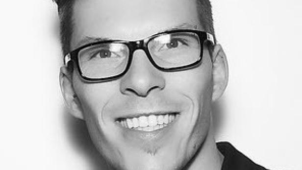 Ein lächelnder Mann mit Brille und Anzug vor einem weißen Hintergrund.