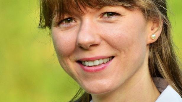 Eine lächelnde Frau mit Perlenkette und dunklem Blazer vor grünem Hintergrund.