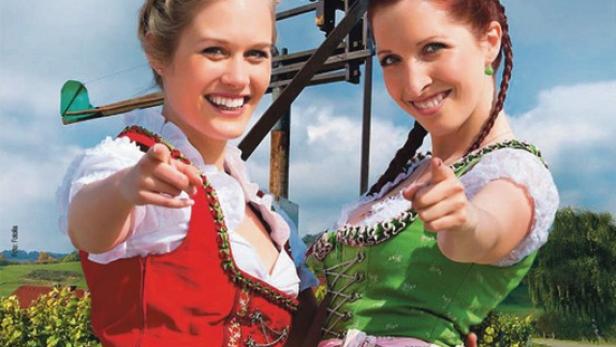 Zwei lächelnde Frauen in Tracht weisen auf den Betrachter vor einer Windmühle in der Steiermark.