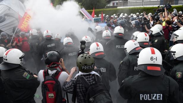Polizisten stehen in einer Reihe inmitten von Rauch, während Demonstranten im Hintergrund zu sehen sind.