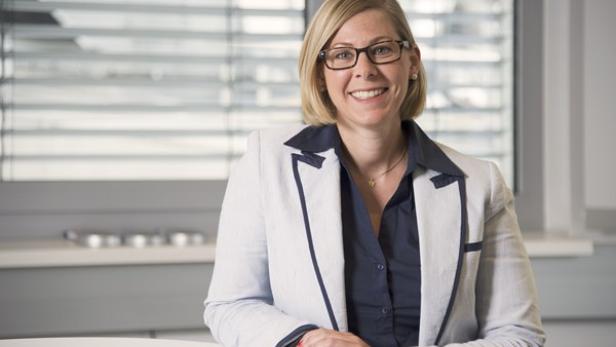 Eine lächelnde Frau mit Brille und Blazer sitzt an einem Tisch.
