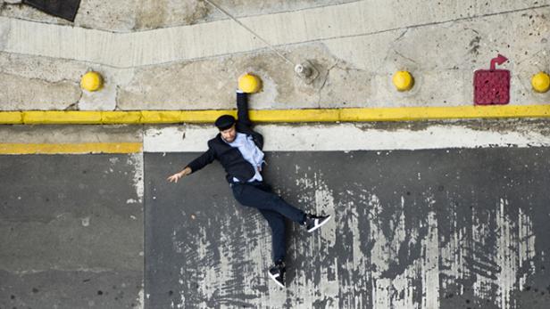 Ein Mann in Anzug und Mütze hängt an einem gelben Knopf unter einer Brücke.