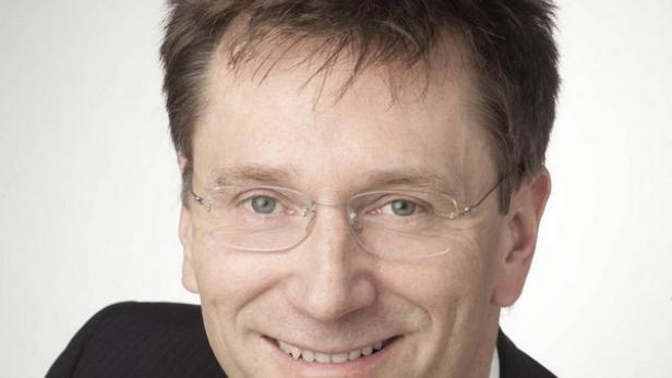 Ein lächelnder Mann mit Brille und Anzug vor einem hellen Hintergrund.