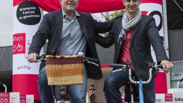 Zwei Männer fahren auf Fahrrädern vor einem „bike2help“-Banner.