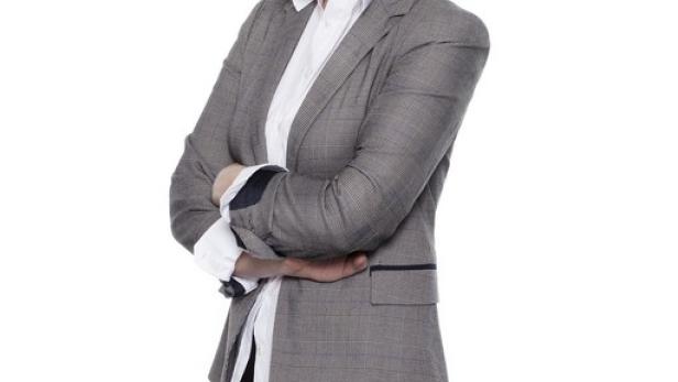 Eine blonde Frau in Lederhose und Blazer lächelt mit verschränkten Armen.