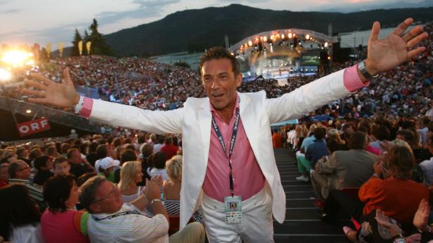 Ein Mann mit weißem Anzug steht vor einer jubelnden Menschenmenge auf einer Open-Air-Veranstaltung.