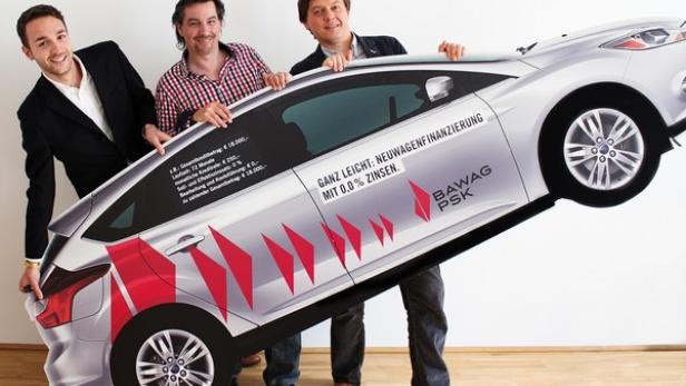 Drei Männer halten eine Pappfigur eines Autos mit Werbung für eine Autofinanzierung der BAWAG P.S.K..