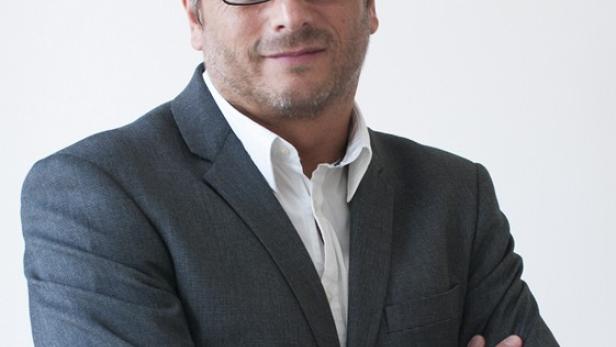 Ein Mann mit Brille und grauem Anzug steht mit verschränkten Armen vor einem weißen Hintergrund.