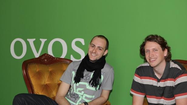 Zwei Männer sitzen vor einem grünen Hintergrund mit dem Schriftzug OVOS.