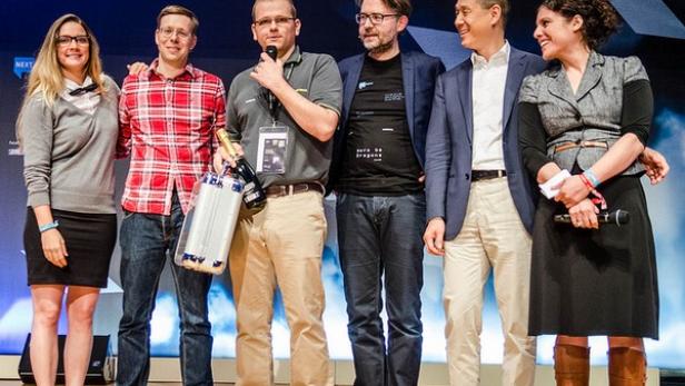 Eine Gruppe von sechs Personen steht auf einer Bühne, wobei eine Person ein Mikrofon hält und eine Flasche Champagner präsentiert.