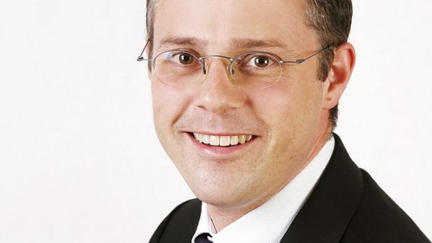 Ein lächelnder Mann mit Brille, Anzug und Krawatte vor einem hellen Hintergrund.