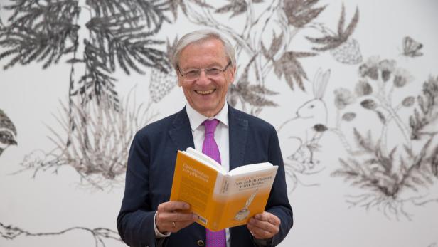 Wolfgang Schüssel lächelt und hält sein Buch „Optimismus verpflichtet“ in die Kamera.