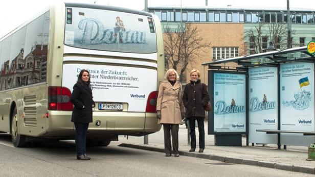 Drei Frauen stehen an einer Bushaltestelle vor einem Bus mit Werbung für die Donau.