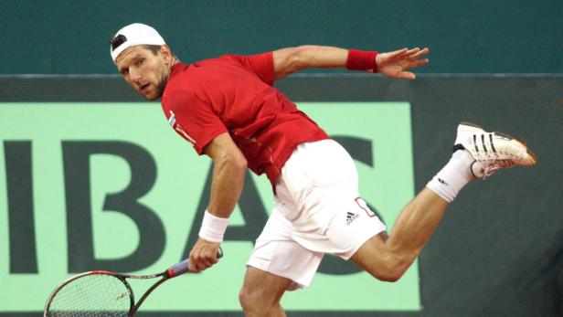 Ein Tennisspieler in rotem Hemd und weißer Hose schlägt einen Ball.